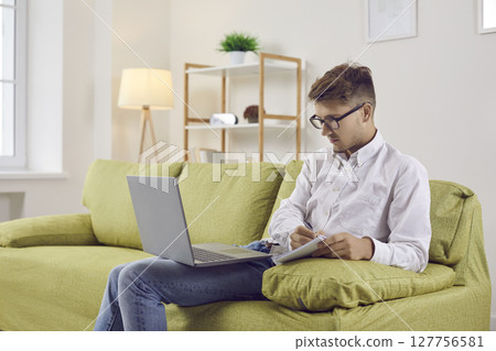 Young man sitting on couch with laptop computer, having online class and taking notes Young man sitting on couch with laptop computer, having online class and taking notes 127756581