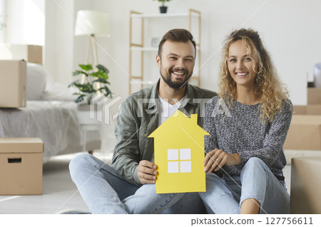 Couple sitting in living room in new apartment, holding paper house and smiling happily 127756611
