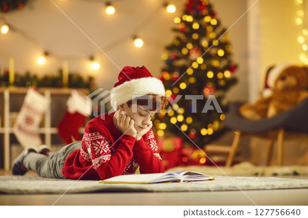 Teen boy in traditional red Christmas clothes lying on floor and reading book at home 127756640