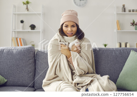 Woman wrapped in a plaid suffering from the cold in a bright living room in the apartment. Woman wrapped in a plaid suffering from the cold in a bright living room in the apartment. 127756654