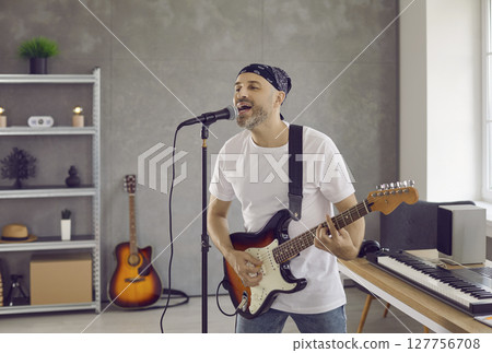 Enthusiastic male singer plays electric guitar and sings into microphone in home studio. Enthusiastic male singer plays electric guitar and sings into microphone in home studio. 127756708
