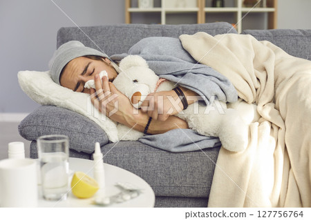 Sick young man lying on couch at home, hugging teddy and blowing nose in paper tissue 127756764