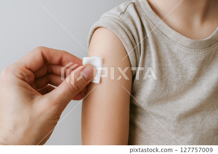 Close up of child arm being cleaned with cotton pad after receiving vaccine, showing care and protection in medical setting. child wears beige shirt Close up of child arm being cleaned with cotton pad after receiving vaccine, showing care and protection in medical setting. child wears beige shirt 127757009
