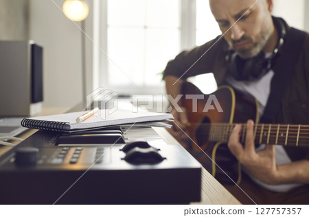 Musician playing guitar and writing music while sitting by keyboard with paper and pencil Musician playing guitar and writing music while sitting by keyboard with paper and pencil 127757357