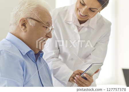 Smiling nurse teaching senior man to use healthcare app or showing photo of his grandchildren Smiling nurse teaching senior man to use healthcare app or showing photo of his grandchildren 127757378
