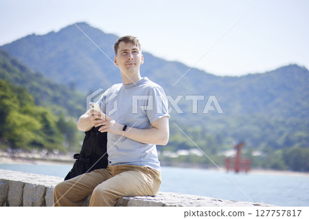 一名男子背著背包，正在使用智慧型手機，背景是大海和鳥居門 127757817
