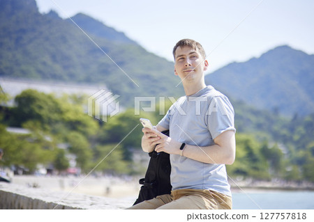 A man with a backpack using a smartphone with the sea and torii gate in the background A man with a backpack using a smartphone with the sea and torii gate in the background 127757818