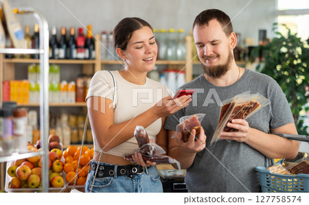 Portrait of man and woman checking the expiration date of dried sausage and jamon using smartphone 127758574