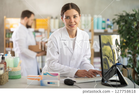 Young woman pharmacist with package of pills, she stands behind the counter near the computer. Pharmacist works in a pharmacy against the background of an male assistant who puts goods on the shelves Young woman pharmacist with package of pills, she stands behind the counter near the computer. Pharmacist works in a pharmacy against the background of an male assistant who puts goods on the shelves 127758618
