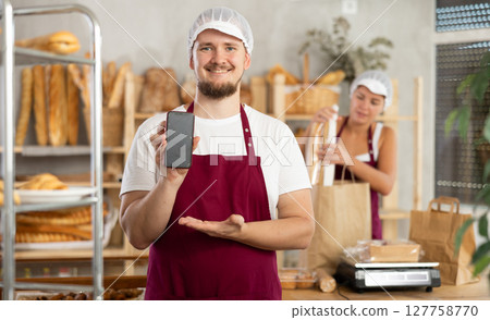 Guy seller in bakery hold out hand with phone, points at screen with finger 127758770