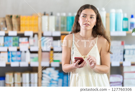 Woman is standing in a pharmacy and looking for goods with a phone in her hands 127760306