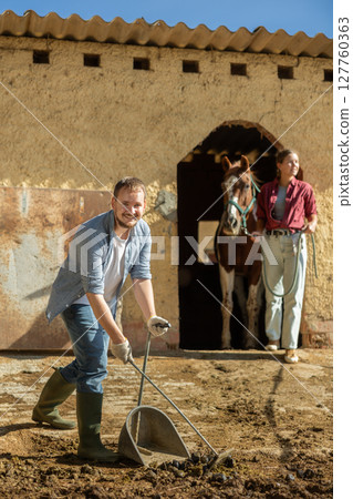 Man cleans up horse poop in horse pasture using broom and dustpan. In background, girl leads horse from stable into paddock Man cleans up horse poop in horse pasture using broom and dustpan. In background, girl leads horse from stable into paddock 127760363