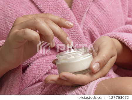 Young woman in pink bathrobe touching cream with finger and holding a cosmetic jar closeup Young woman in pink bathrobe touching cream with finger and holding a cosmetic jar closeup 127763072