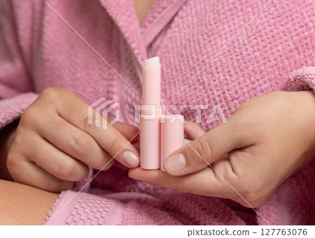Woman in pink bathrobe holding in hand opened lip balm stick with a lid closeup, girly mockup 127763076