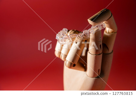 Wooden hand of an impersonal mannequin holds transparent polymer dental aligners on a colored background for teeth alignment. Aesthetic and comfortable teeth alignment 127763815