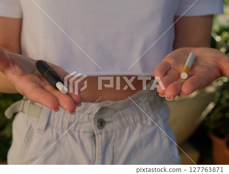 The girl holds a cigarette and an electronic cigarette in her hands. Choice of which devices to smoke. The girl holds a cigarette and an electronic cigarette in her hands. Choice of which devices to smoke. 127763871
