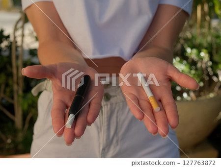 The girl holds a cigarette and an electronic cigarette in her hands. Choice of which devices to smoke. The girl holds a cigarette and an electronic cigarette in her hands. Choice of which devices to smoke. 127763872