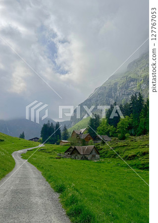 Axalp region of Bern canton Swiss rural landscape winds through lush green grass, guiding toward cozy cabins nestled against majestic mountains. 127767053
