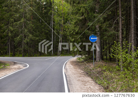 An empty asphalt road winding through a green forest, traffic sign directing left An empty asphalt road winding through a green forest, traffic sign directing left 127767630