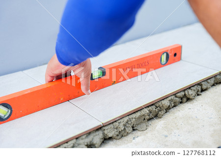 A hand carefully uses an orange level to check the alignment of a tile placed over gray mortar, ensuring precision in tile installation for a flawless finish. A hand carefully uses an orange level to check the alignment of a tile placed over gray mortar, ensuring precision in tile installation for a flawless finish. 127768112