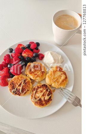 Homemade syrniki with berries and sour cream served on a white plate with a cup of coffee. Concept of cozy morning, balanced breakfast, indulgence and homemade comfort 127768813