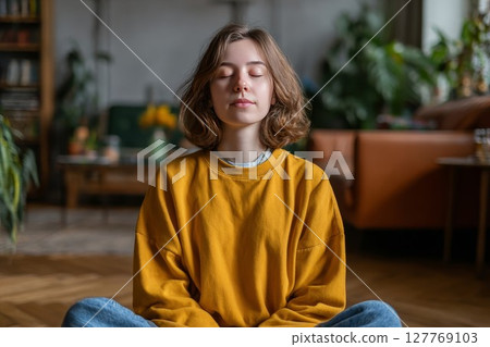 European woman in yellow sweater meditating indoors. Ideal for stress relief or breathing tips. Adds tranquil, uplifting vibe to health posts. 127769103
