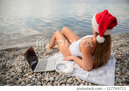 A woman is laying on the beach with a laptop and headphones on her head. She is wearing a red Santa hat. 127769712