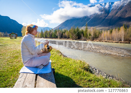 Woman playing a Tibetan singing bowl by a mountain river in Altai. Concept of sound healing, spiritual alignment, and deep connection with natural elements 127770863