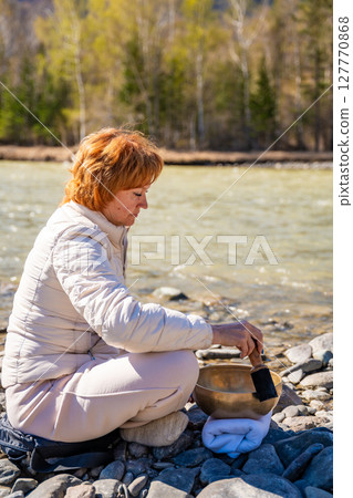 Woman performing a sound ritual with a Tibetan singing bowl by the Altai river. Concept of feminine energy, sacred vibration, and shamanic connection to the Earth 127770868