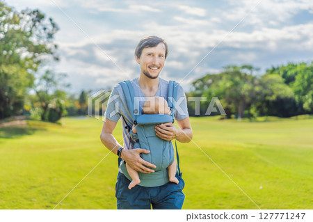Young father walks in nature with his baby son nestled close in a parent-facing ergonomic baby carrier. Bonding, warmth, and modern parenting concept in a peaceful outdoor setting 127771247