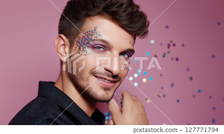 Smiling young man with glitter makeup on pink background posing with confident expression. Studio portrait with colorful sparkles. LGBT pride, queer identity, and self-expression concept. Design for Smiling young man with glitter makeup on pink background posing with confident expression. Studio portrait with colorful sparkles. LGBT pride, queer identity, and self-expression concept. Design for 127771794