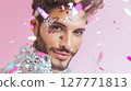 Close-up portrait of young man with glitter makeup and sequin outfit smiling under falling confetti on pink background. Studio shot with soft lighting. LGBT pride, queer identity, and self-expression 127771813