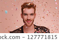 Smiling young man wearing sequin jacket posing under falling confetti on peach background. Studio portrait with festive atmosphere. Celebration, pride, and LGBT identity concept. Design for poster 127771815