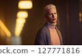 Stylish young man with platinum blonde hair wearing turtleneck sweater and shawl standing in dimly lit hallway with warm lights. Fashion portrait with modern identity and soft atmospheric lighting 127771816