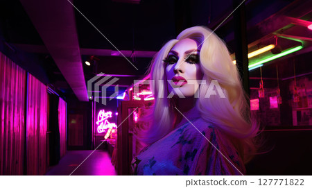 Drag queen with glamorous makeup and long blonde wig posing in neon-lit hallway of night club. Studio portrait with colorful lighting. Drag performance and LGBT concept. Design for poster, banner 127771822