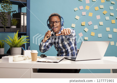 Black businessman using mobile device as a microphone, looking at camera and performing an audio session. Happy male entrepreneur seated at office desk, wearing headphones and recording a song. Black businessman using mobile device as a microphone, looking at camera and performing an audio session. Happy male entrepreneur seated at office desk, wearing headphones and recording a song. 127771901