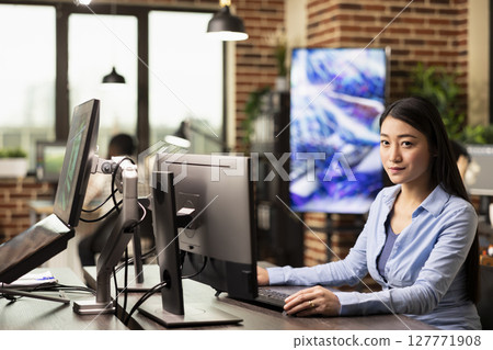 Portrait of asian woman sitting and working on her desktop pc in brick wall workplace. Young female office worker at office desk, using her computer and looking at camera in creative agency company. Portrait of asian woman sitting and working on her desktop pc in brick wall workplace. Young female office worker at office desk, using her computer and looking at camera in creative agency company. 127771908