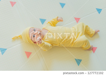 Adorable baby in a yellow hat lying on a play mat. Cozy newborn moment with soft textures and bright colors. Infant development, sensory play, and early childhood concept Adorable baby in a yellow hat lying on a play mat. Cozy newborn moment with soft textures and bright colors. Infant development, sensory play, and early childhood concept 127772102