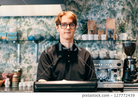 Young waiter serving customer at cash point in cafe. Man working with POS terminal. Cashier, barista checking for payment receipt. Hospitality, server and preparing a slip at the till in coffee shop. 127772439