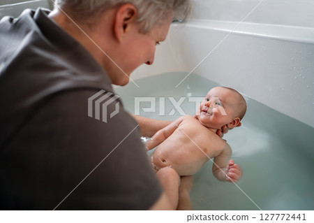 Top view smiling baby bathing and swimming in the bath. Baby learning to swim and playing sports. Father enjoys bath time with his curious baby in a cozy bathroom. Fatherhood concept. Selective focus. Top view smiling baby bathing and swimming in the bath. Baby learning to swim and playing sports. Father enjoys bath time with his curious baby in a cozy bathroom. Fatherhood concept. Selective focus. 127772441