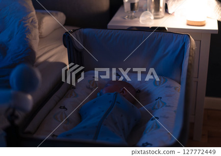 A baby wrapped snugly in cocoon swaddle bag is peacefully sleeping in a crib near parent bed with baby video nanny monitor camera. Safety newborn baby sleep. Modern technology. Soft selective focus A baby wrapped snugly in cocoon swaddle bag is peacefully sleeping in a crib near parent bed with baby video nanny monitor camera. Safety newborn baby sleep. Modern technology. Soft selective focus 127772449