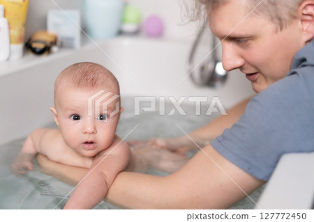 Cute baby bathing and swimming in the bath. Baby learning to swim and playing sports. Father enjoys bath time with his curious baby in a cozy bathroom. Fatherhood concept. Selective focus. 127772450