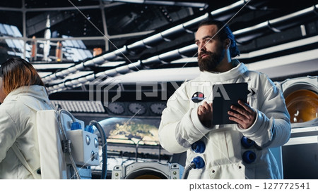 Astronaut reading mission plan on tablet before stepping out for scheduled operations outside space station. Cosmonaut checks analytics on device, preparing to leave after landing on planet Astronaut reading mission plan on tablet before stepping out for scheduled operations outside space station. Cosmonaut checks analytics on device, preparing to leave after landing on planet 127772541