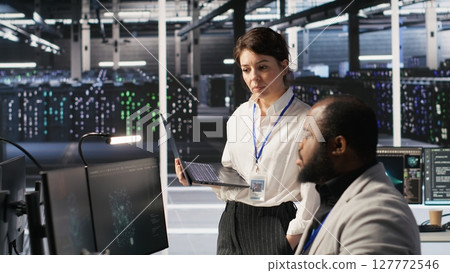 Happy data center manager providing positive feedback to engineer developing neural networks software on computer. Cheerful supervisor pleased by employee work implementing AI in server room, camera A 127772546
