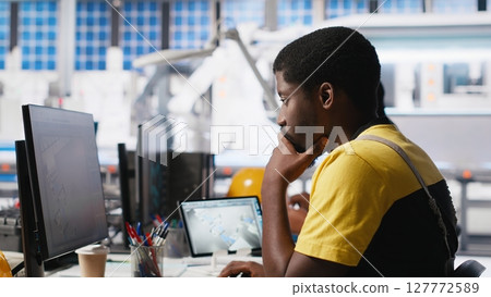 Black worker programming robotic assembly line in solar cell factory, working with high tech software for automation. Male engineer testing photovoltaic system, machinery. Camera A. Black worker programming robotic assembly line in solar cell factory, working with high tech software for automation. Male engineer testing photovoltaic system, machinery. Camera A. 127772589