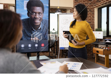 African american man on digital monitor, having financial discussion via video call with businesswomen in startup office. Asian manager standing with tablet, presenting weekly reports to her coworkers 127772592