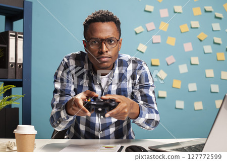 Black male professional at his desk, holding a gaming controller and having fun playing video games during a break at work. African american marketing specialist fully engaged in a gaming session. Black male professional at his desk, holding a gaming controller and having fun playing video games during a break at work. African american marketing specialist fully engaged in a gaming session. 127772599