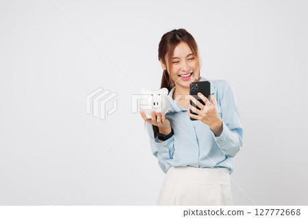 Happy Asian woman holding piggy bank and phone on hand showing bank features planning money saving, mobile invest, and secure bank finance strategy. isolated studio white background 127772668