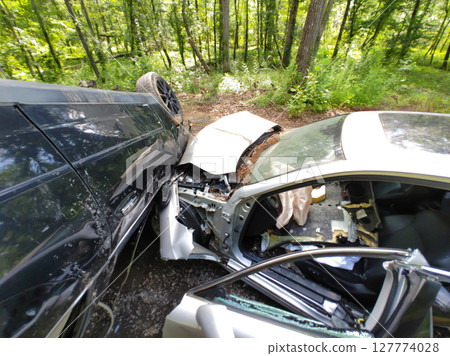 Two cars are heavily damaged after collision on secluded forest road amid dense greenery trees. 127774028