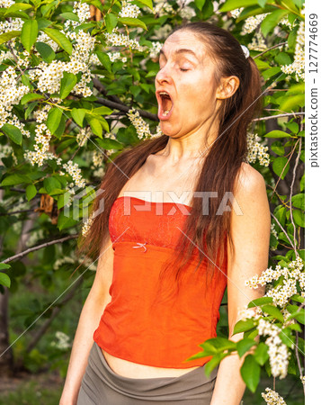 Young woman sneezes in the park against the background of a flowering tree. Allergy to pollen concept. 127774669